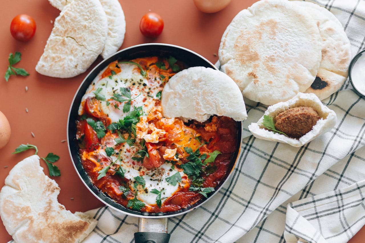 Top view of shakshuka meal with pita bread, cherry tomatoes, and fresh herbs.