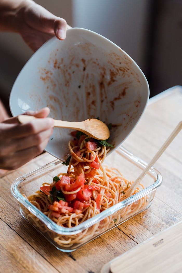 Hands transferring spaghetti with tomato sauce into a glass container using a wooden spoon.
